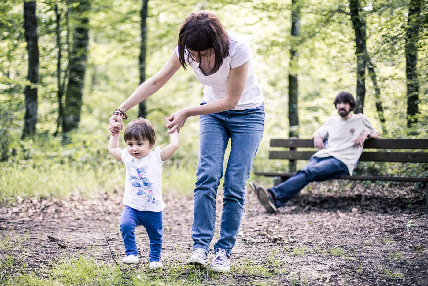 Une petite fille apprend à marcher en forêt aidée de sa maman et sous le regard attentif de son papa